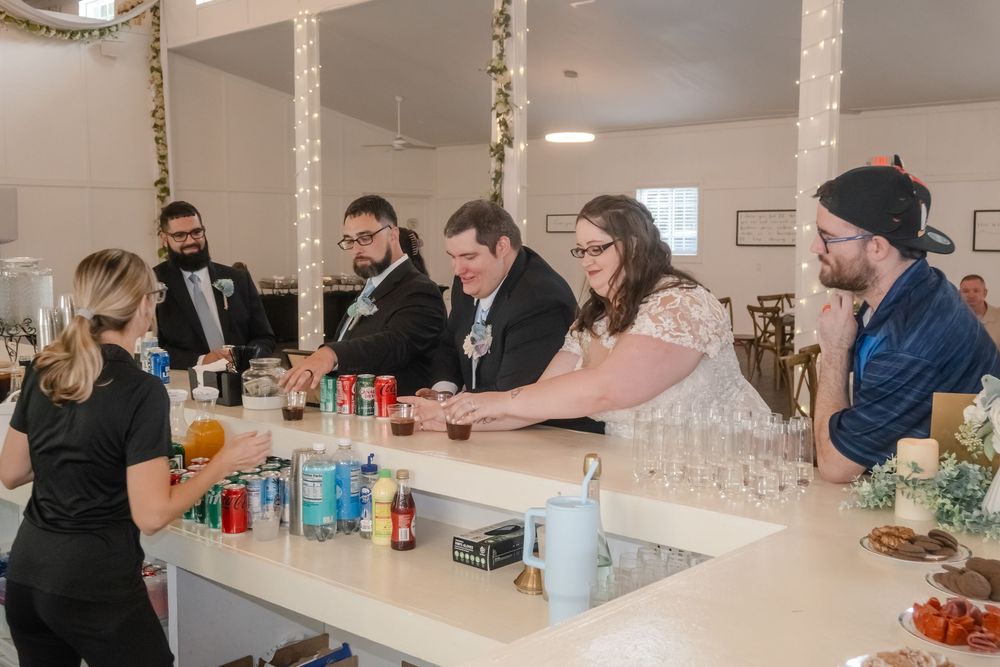 Wedding guests and couple gather at a decorated bar venue, with drinks and glassware displayed on the white counter.