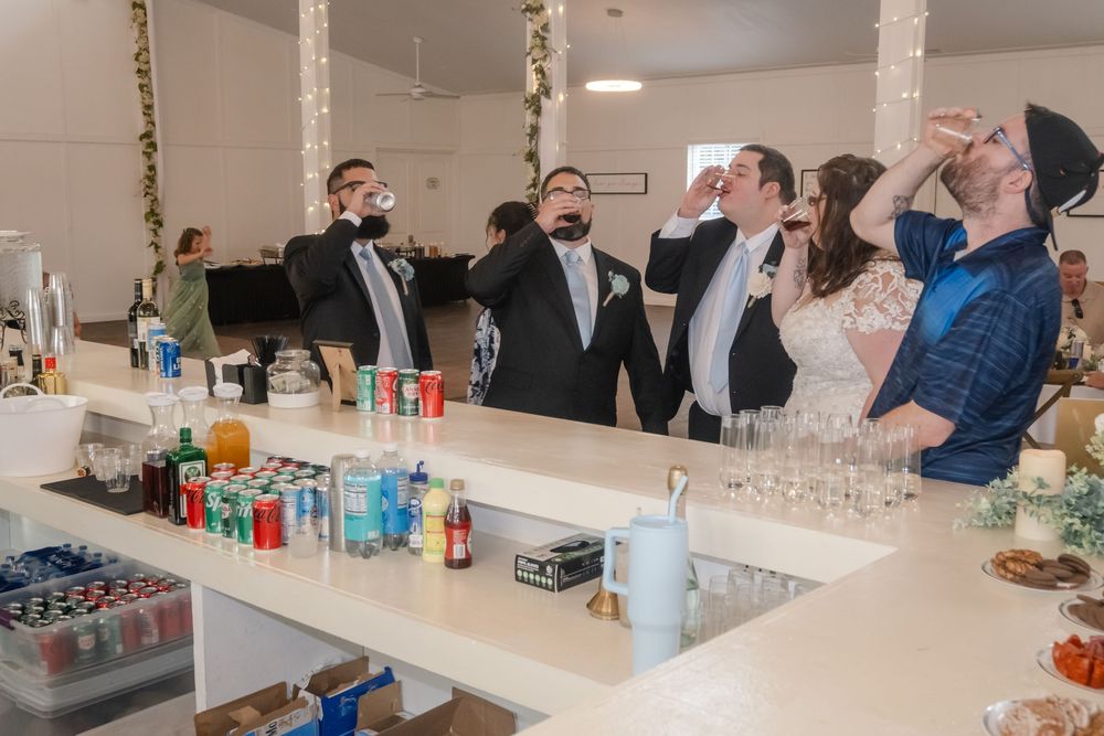 Wedding party doing shots at a white bar during reception celebration with drinks and glassware displayed.
