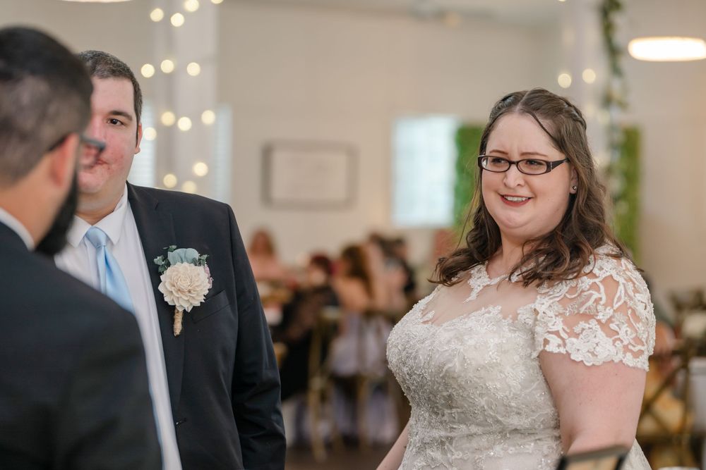 Bride in lace wedding dress and groom in suit smiling during indoor wedding ceremony with bokeh lights.