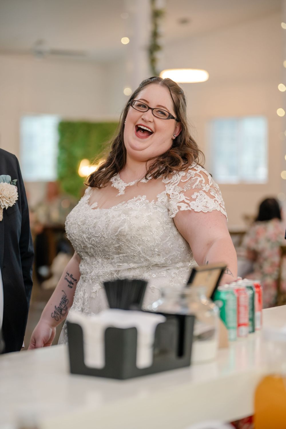 Laughing bride in lace wedding gown with glasses at wedding reception bar.