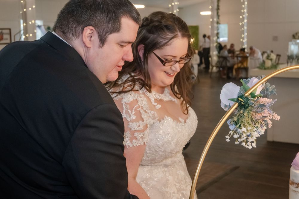 Bride in lace gown and groom in black suit signing wedding documents at indoor ceremony with floral hoop decoration.