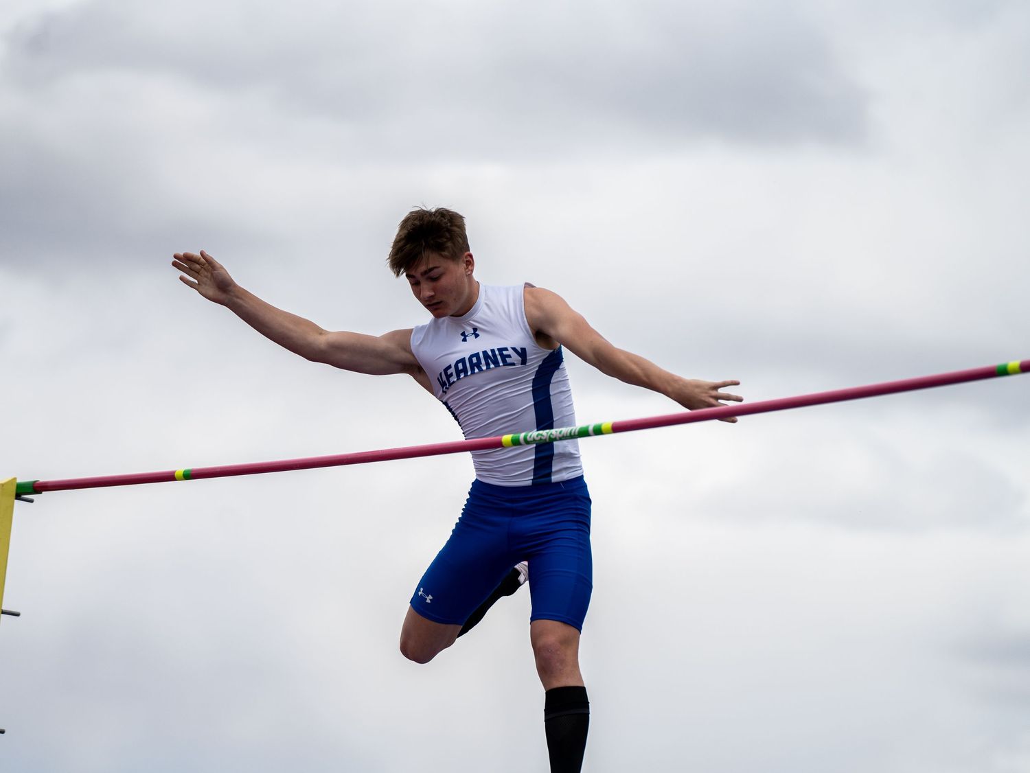 Athlete attempts pole vault clearance against cloudy sky.