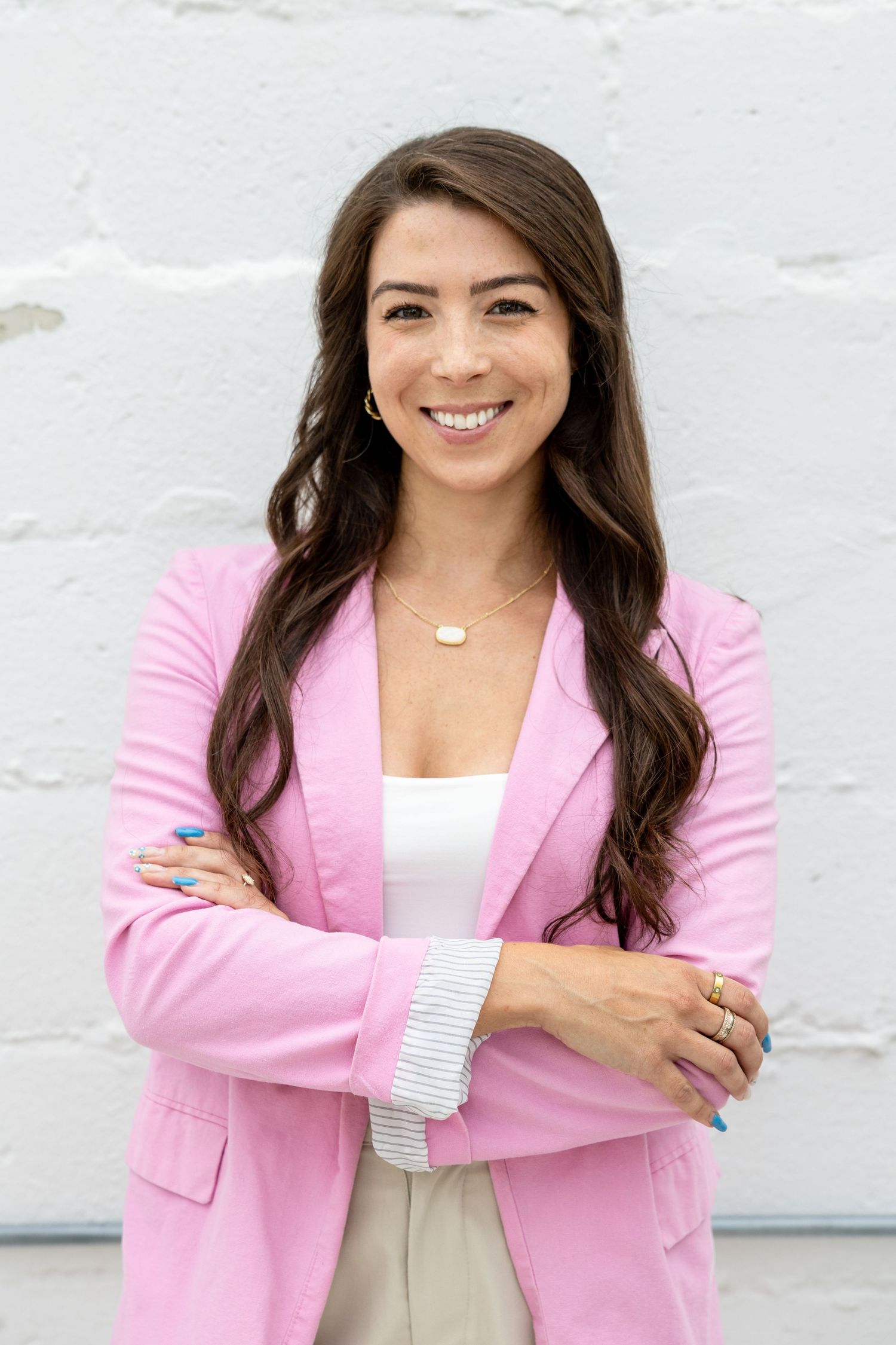 Confident brunette woman in pink blazer smiling with arms crossed against white brick wall.