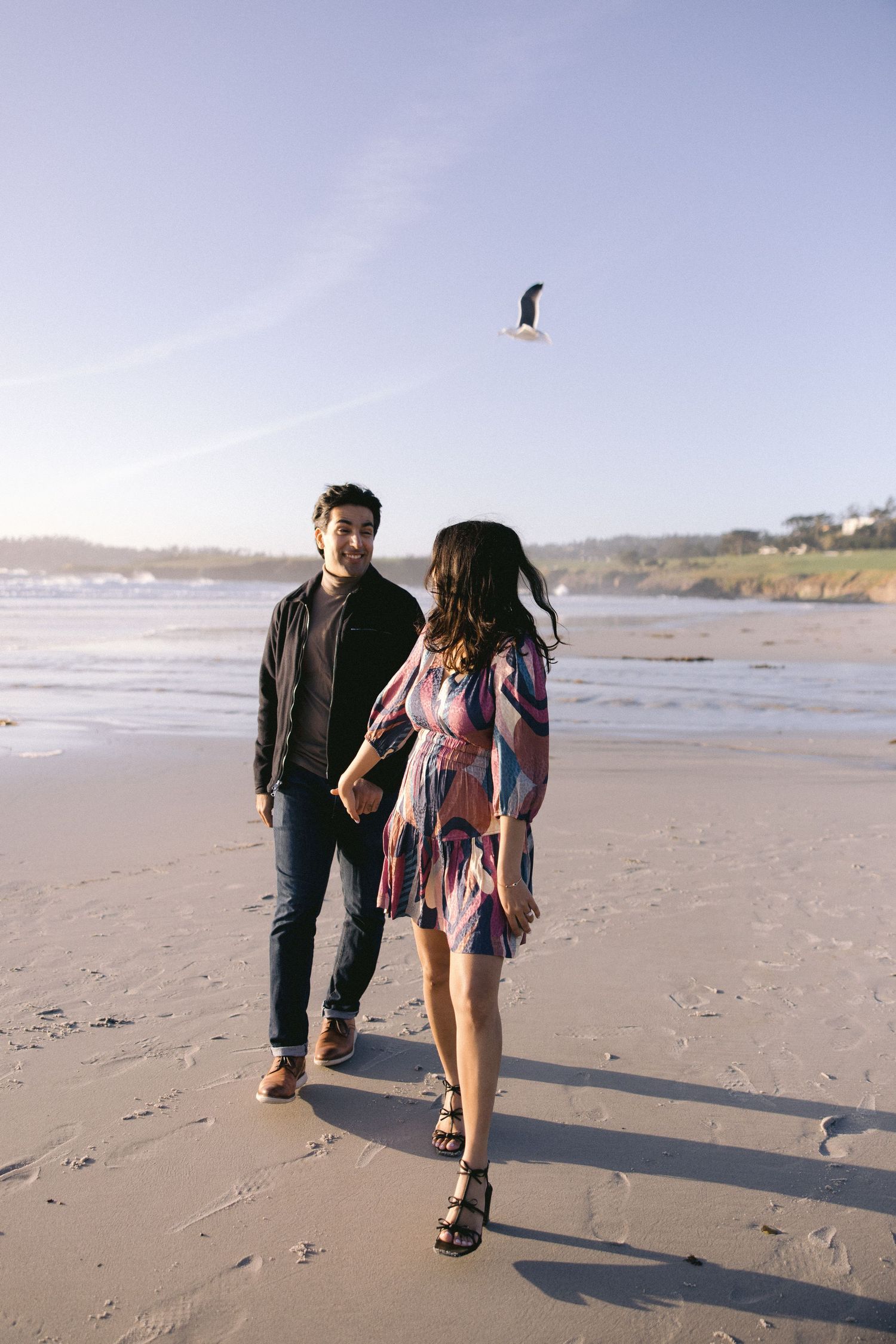 A couple walks along a scenic beach shoreline at sunset while a seagull soars overhead.
