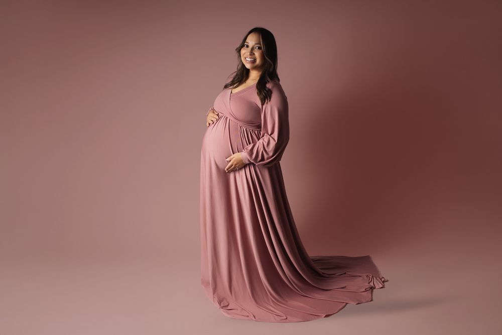 A person in a flowing mauve gown poses gracefully in a series of maternity portraits against a rosy backdrop.