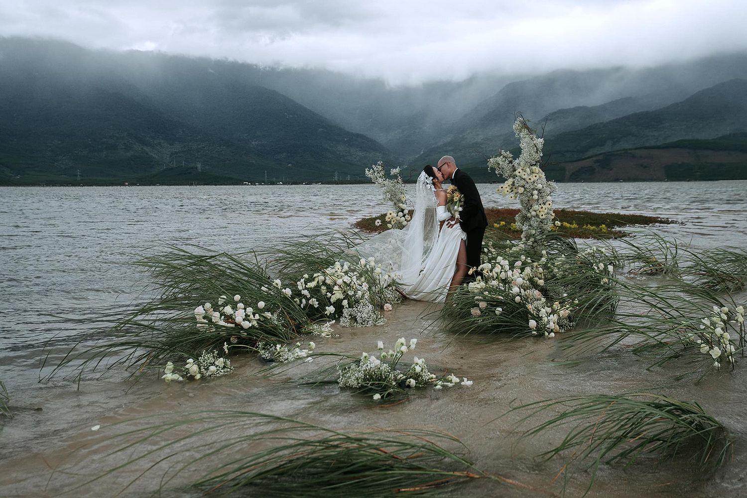Romantic lakeside wedding setup with floral arrangements against misty mountain backdrop.
