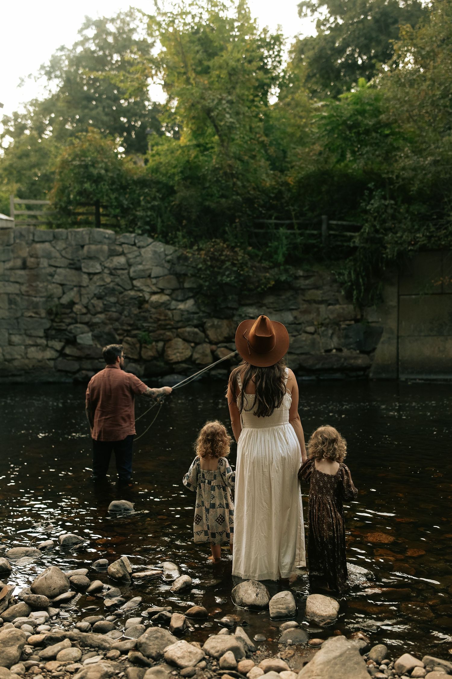 A family stands on rocks by a serene pond with stone walls and lush greenery in the background.