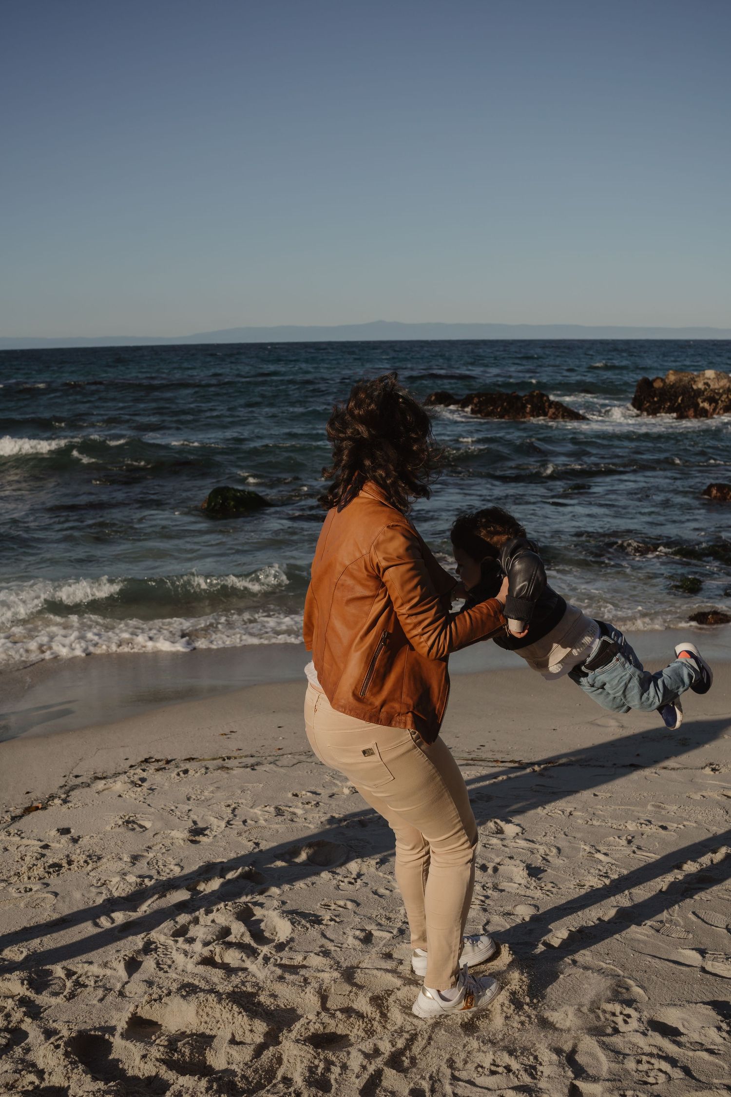 Person in orange jacket plays with small dog on rocky beach shore at sunset while waves crash against rocks in background.