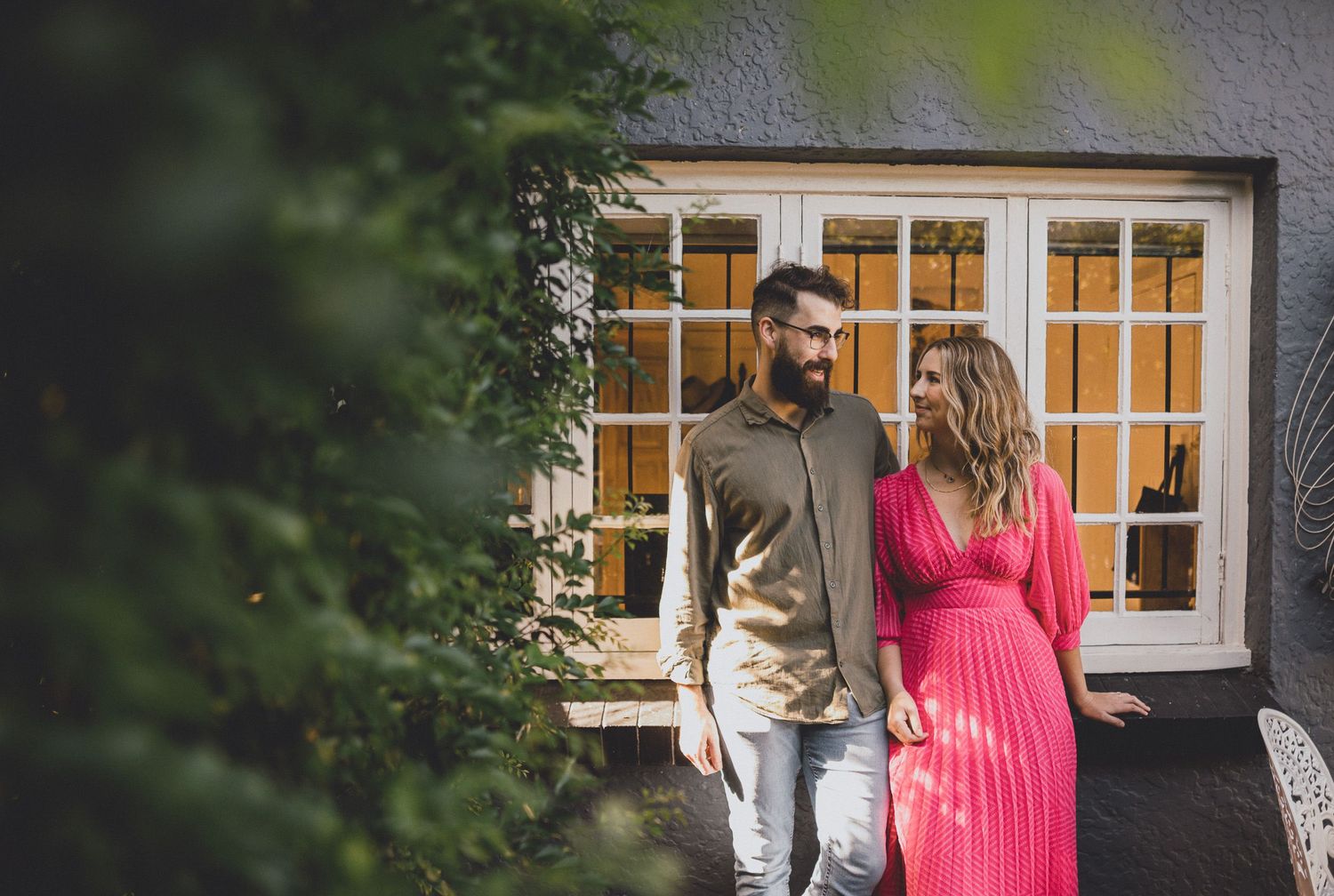 Couple standing by a cottage window wearing pink dress and casual attire.