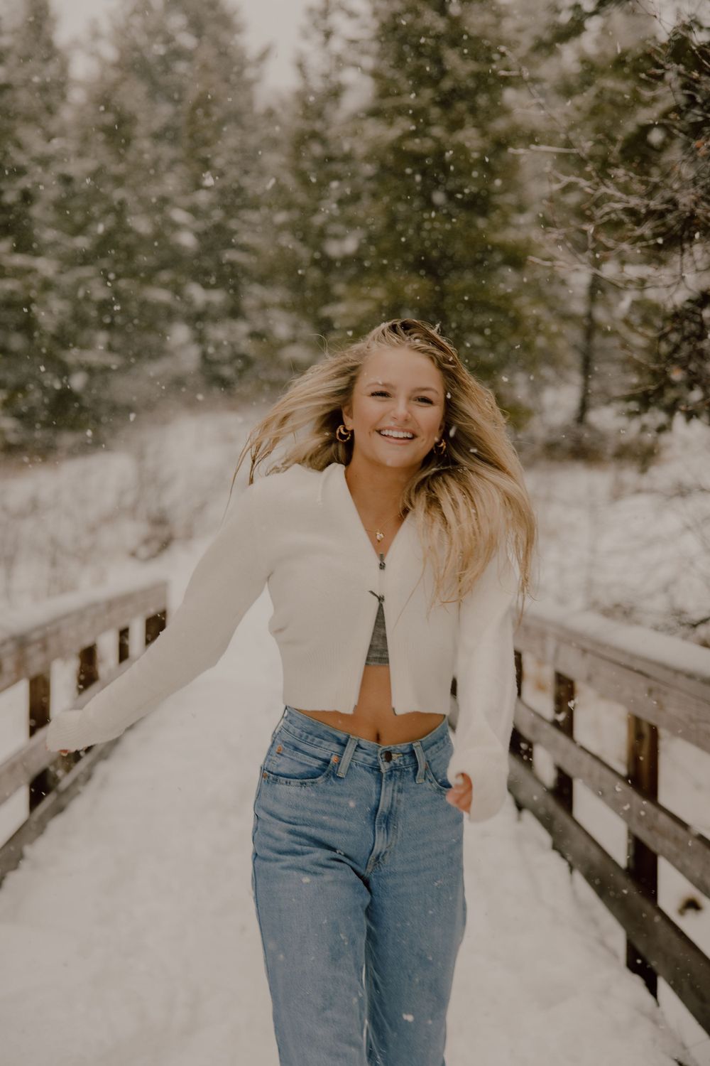 Person wearing white crop top and jeans poses on snowy wooden bridge with evergreen trees in background.