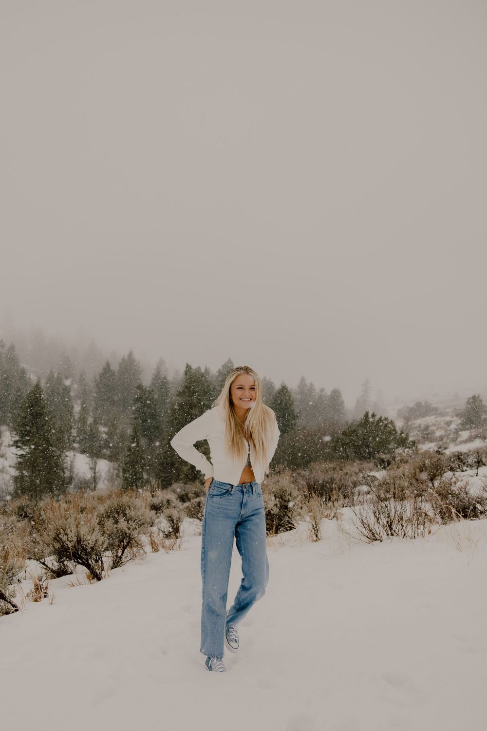 A woman in a white sweater and jeans poses in a snowy winter landscape with evergreen trees in the foggy background.