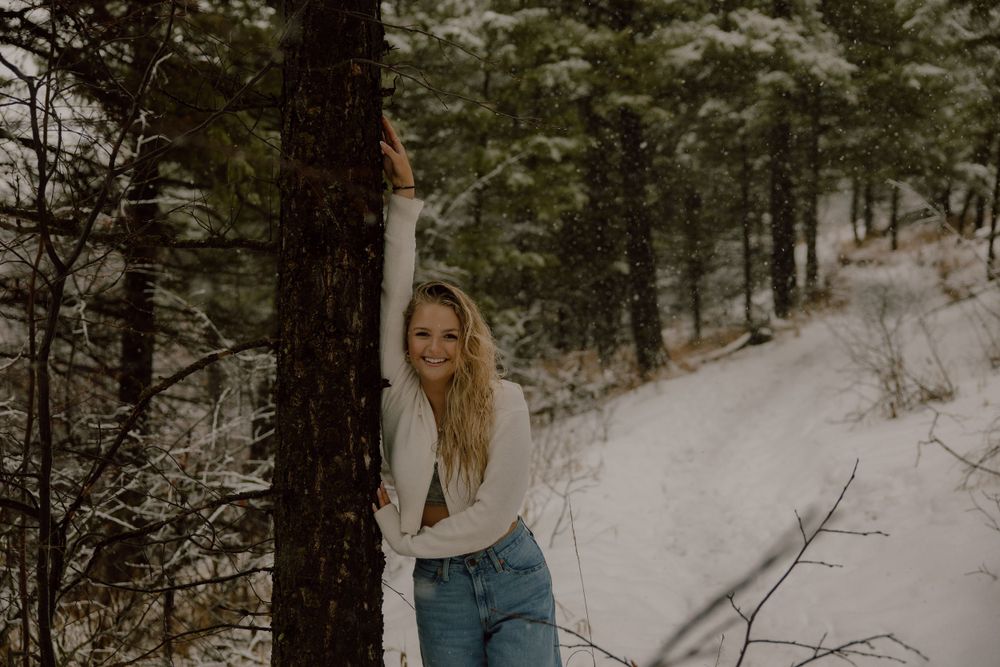 Someone in casual denim stands against a tree trunk in a snowy winter forest setting.