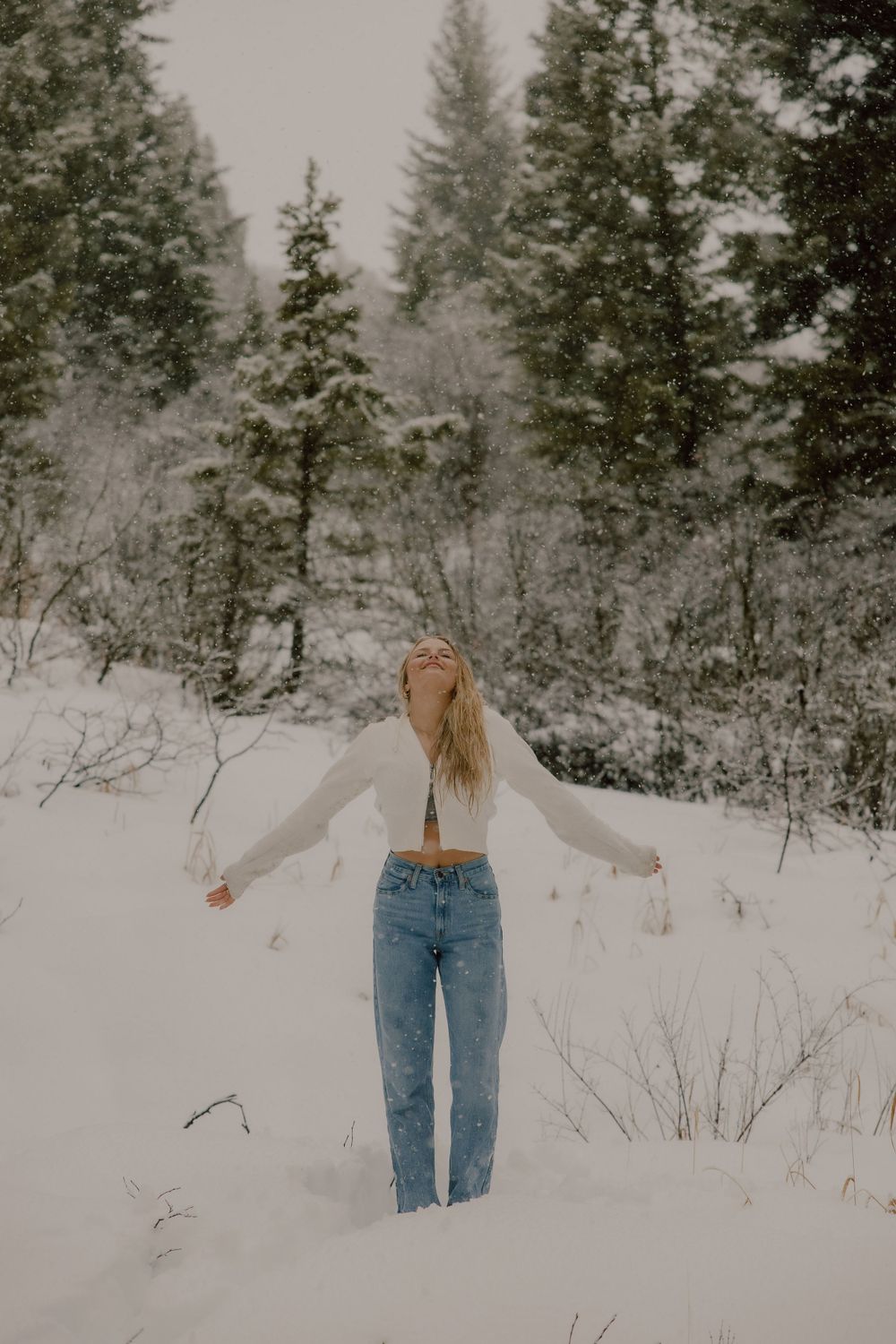 A person in jeans and white top stands with arms outstretched in a snowy winter forest landscape.