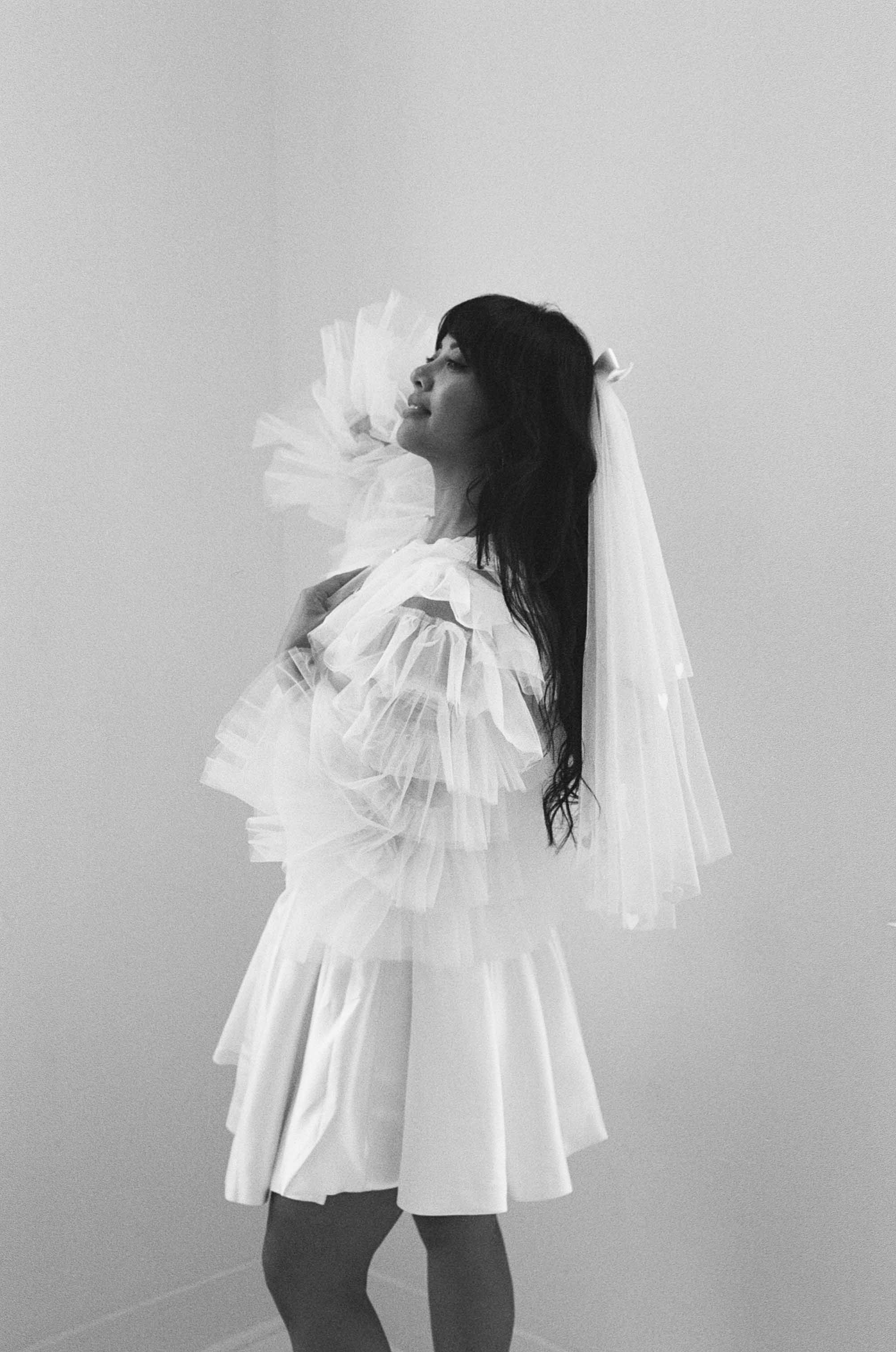 Black and white fashion photo featuring ruffled white dress and bridal veil in artistic pose.