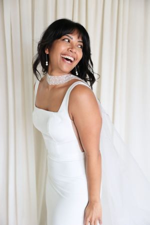 A bride in a sleek white dress and lace choker laughs joyfully against a light backdrop.