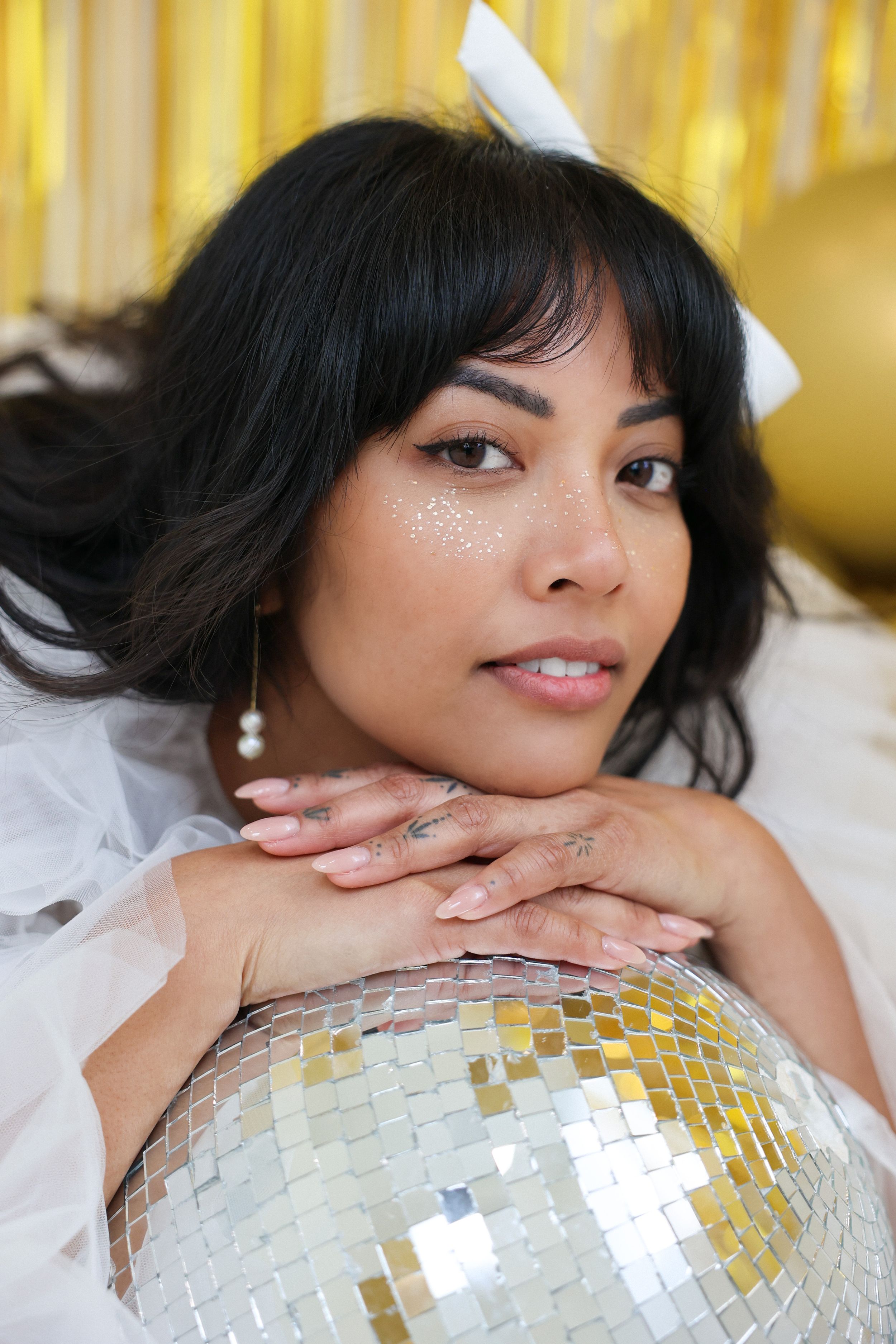 Glamorous disco ball photoshoot with sparkly makeup and white hair bow against gold tinsel background.