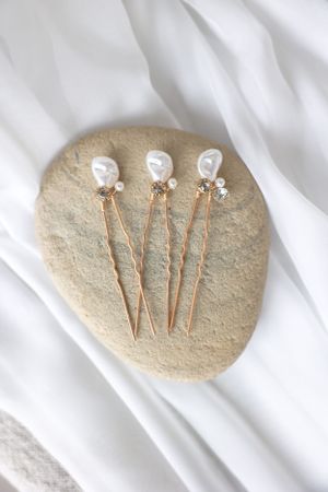 Three delicate pearl hair pins with gold stems displayed on a stone against white fabric.