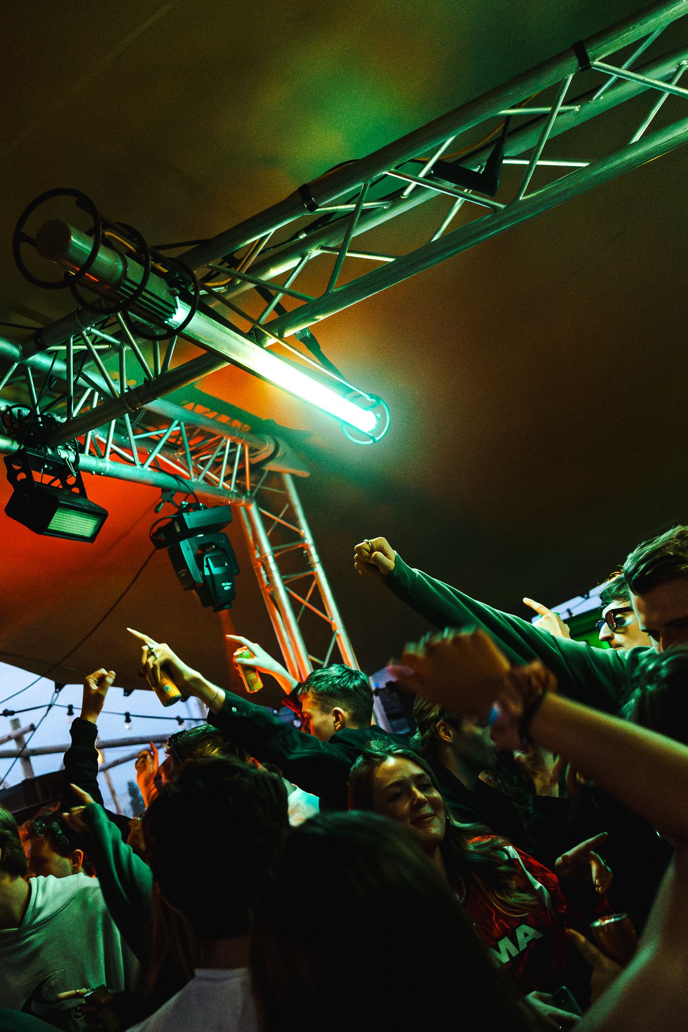 Concert stage lighting truss with green and orange illumination over an energetic crowd at a live music performance.