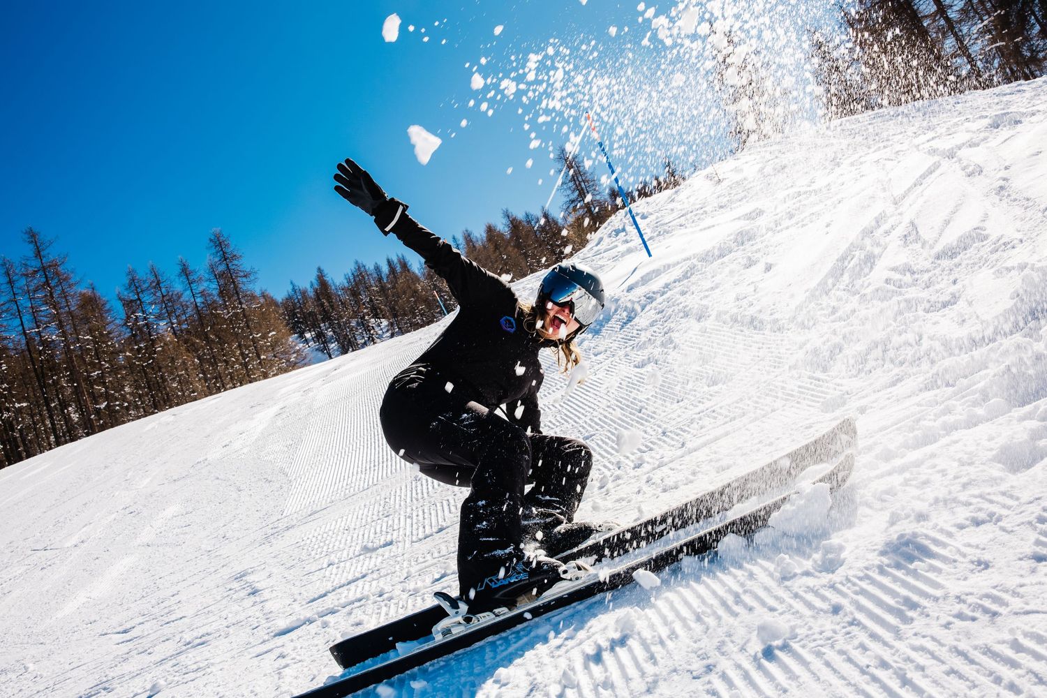 Snowboarder in black gear carving down a snowy slope on a sunny day with blue skies and snow spray.