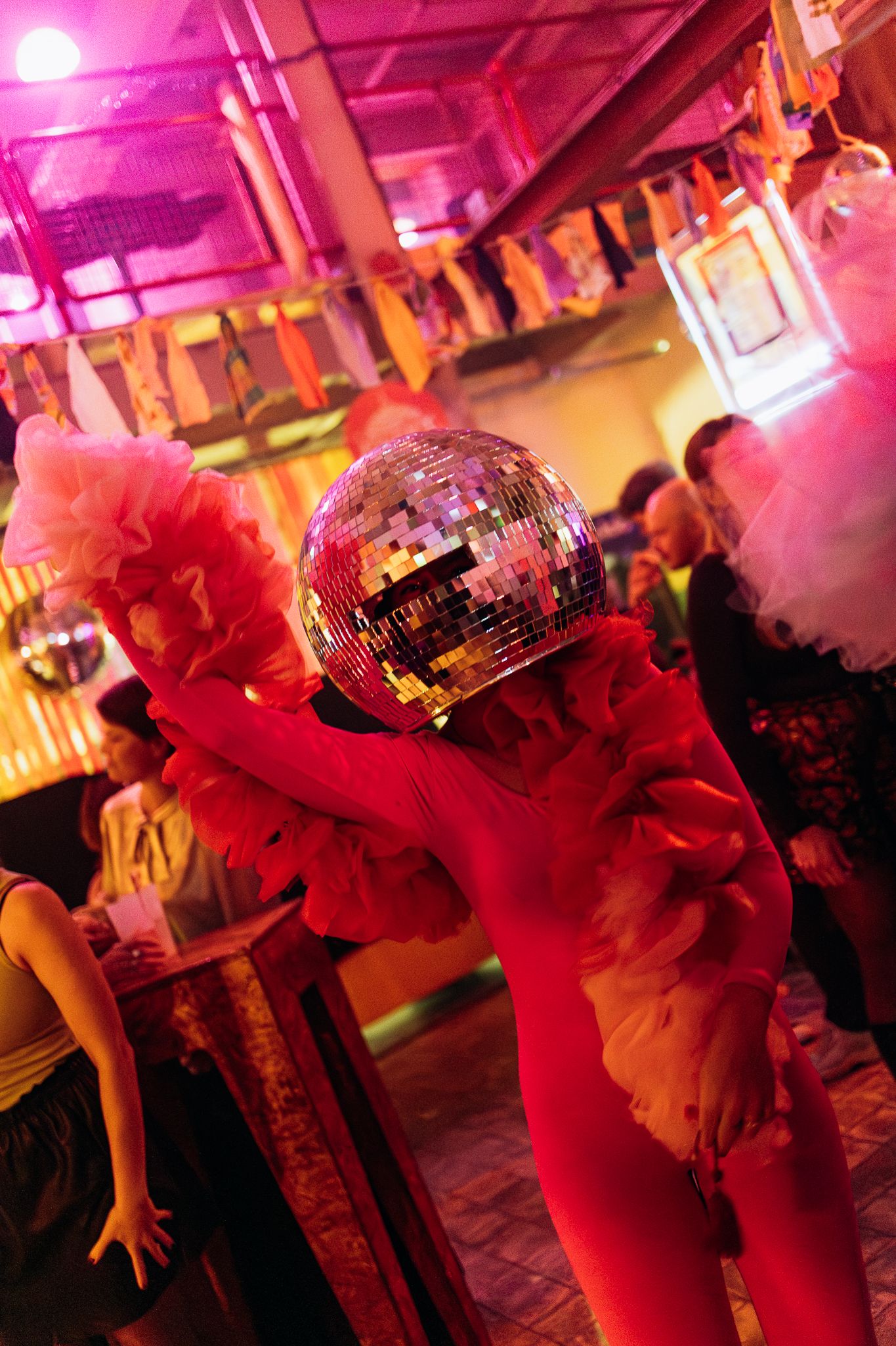 A glittering disco ball hangs above a nightclub dance floor bathed in vibrant pink and red lighting.