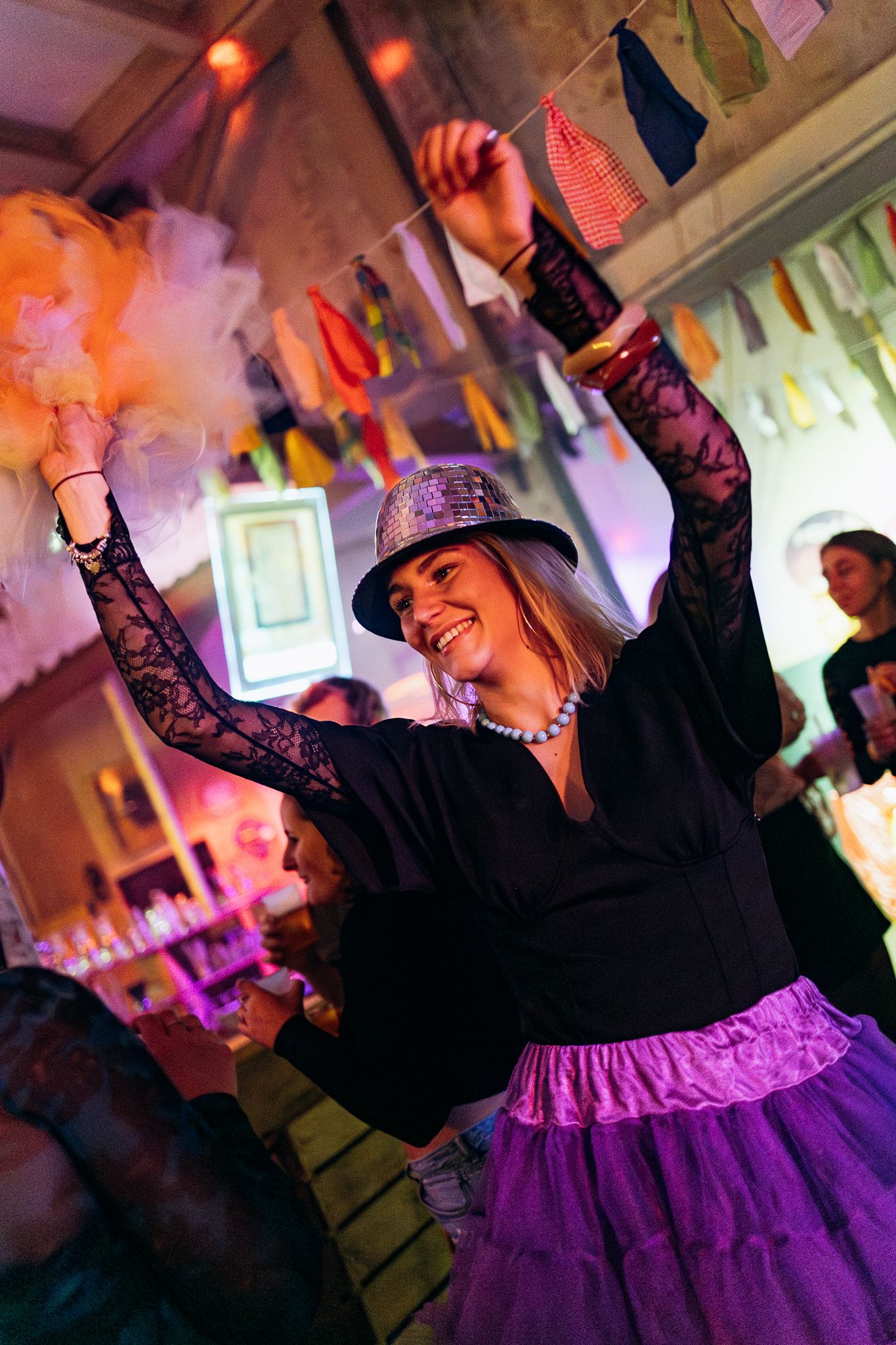 A dancer in a black top and purple tutu skirt performs energetic dance moves in a dimly lit room with colorful lighting.