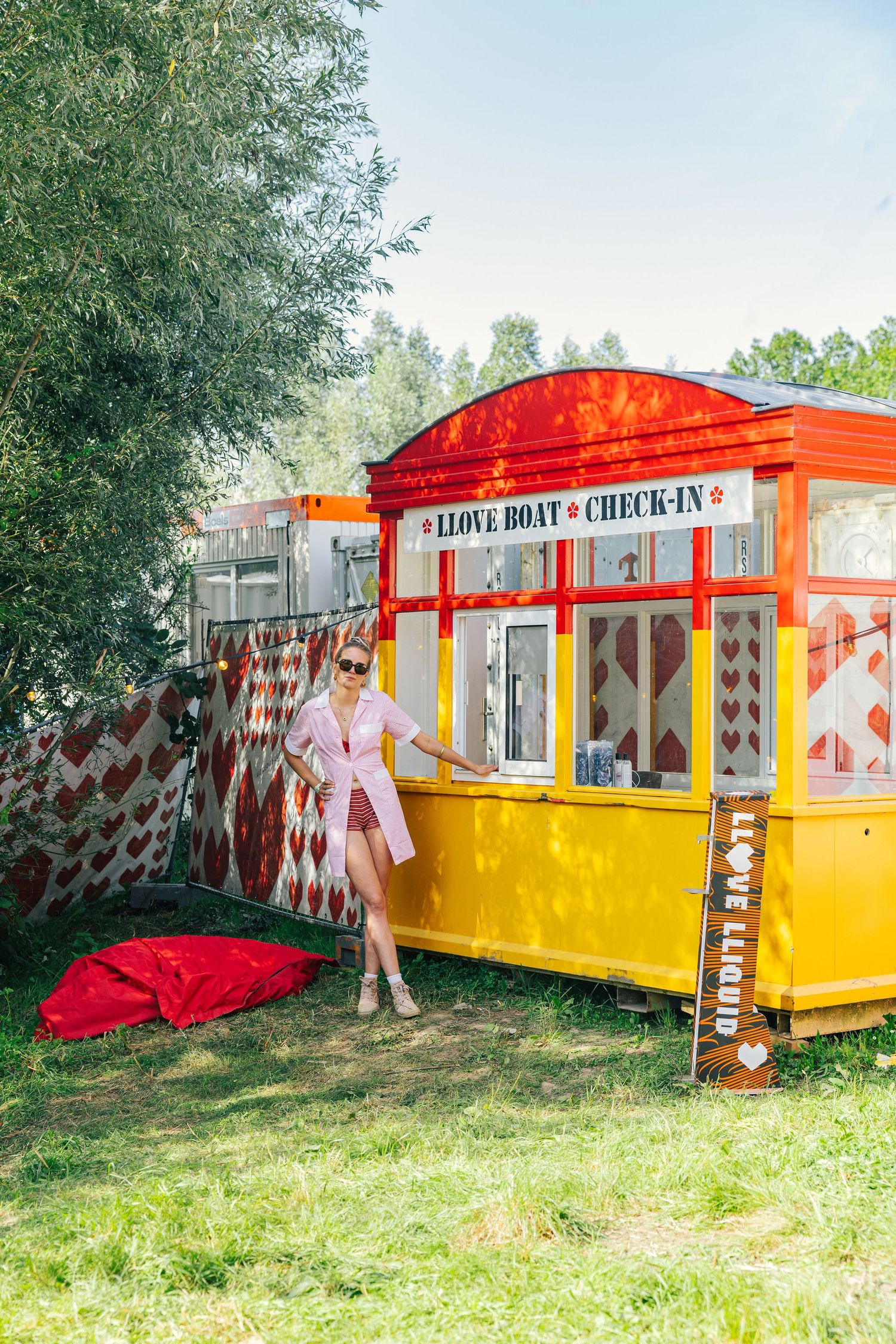 Woman in pink dress posing by colorful yellow and red Llove Boat Check-In booth at outdoor festival.