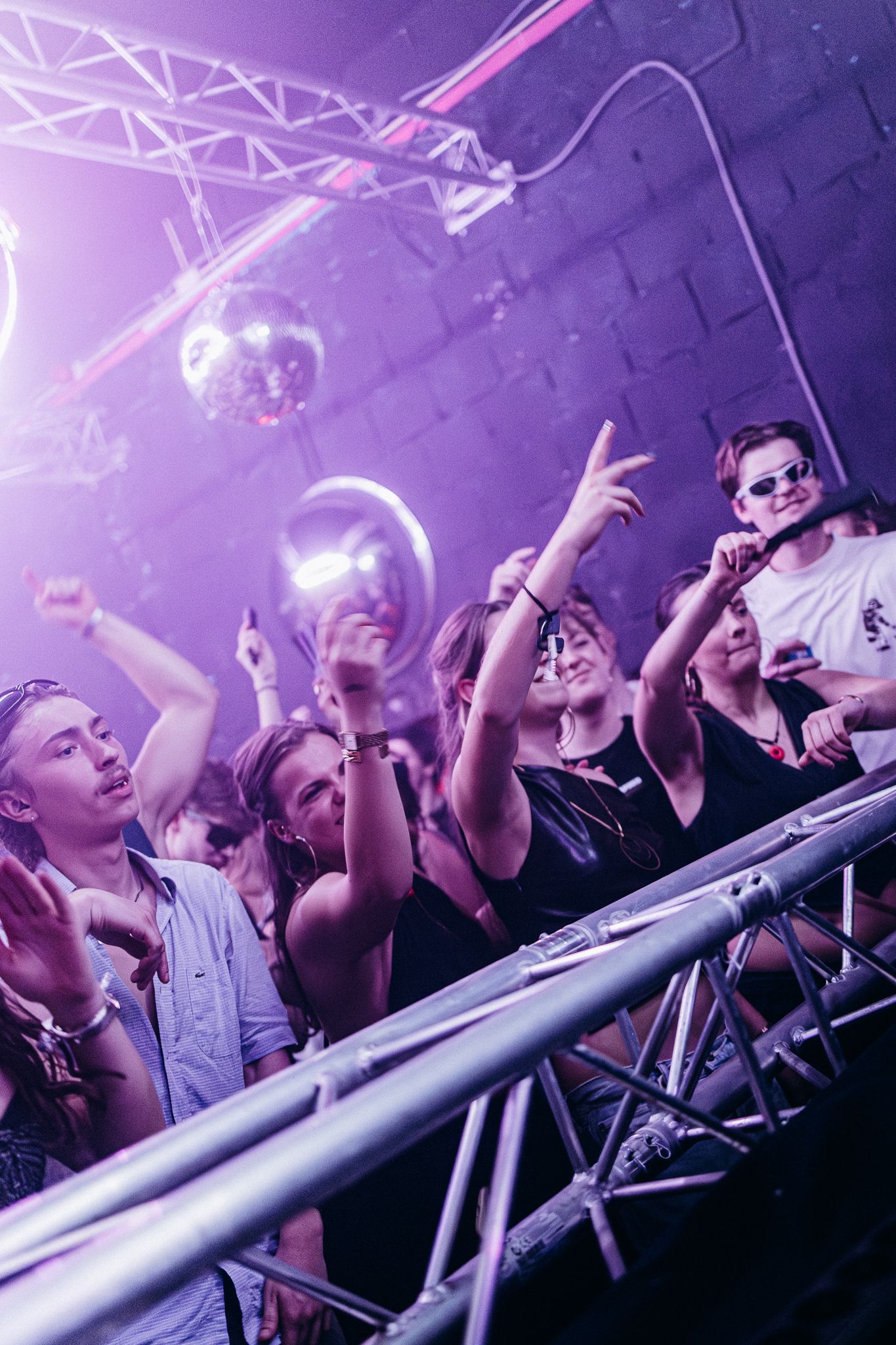 Concert crowd cheering at a music venue with purple lighting and metal barriers in the foreground.