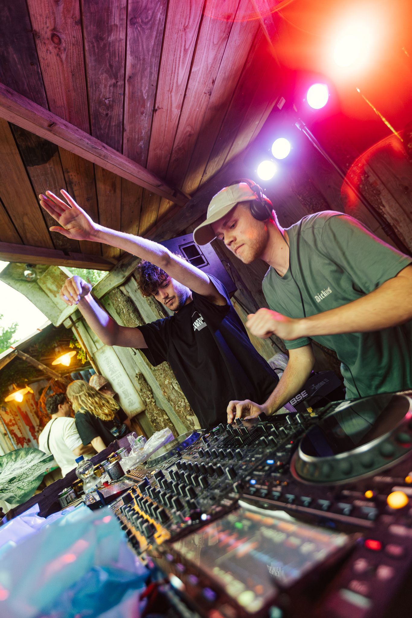 DJ controls mixer equipment under colorful red and blue lighting at a party venue with wooden walls and ceiling.