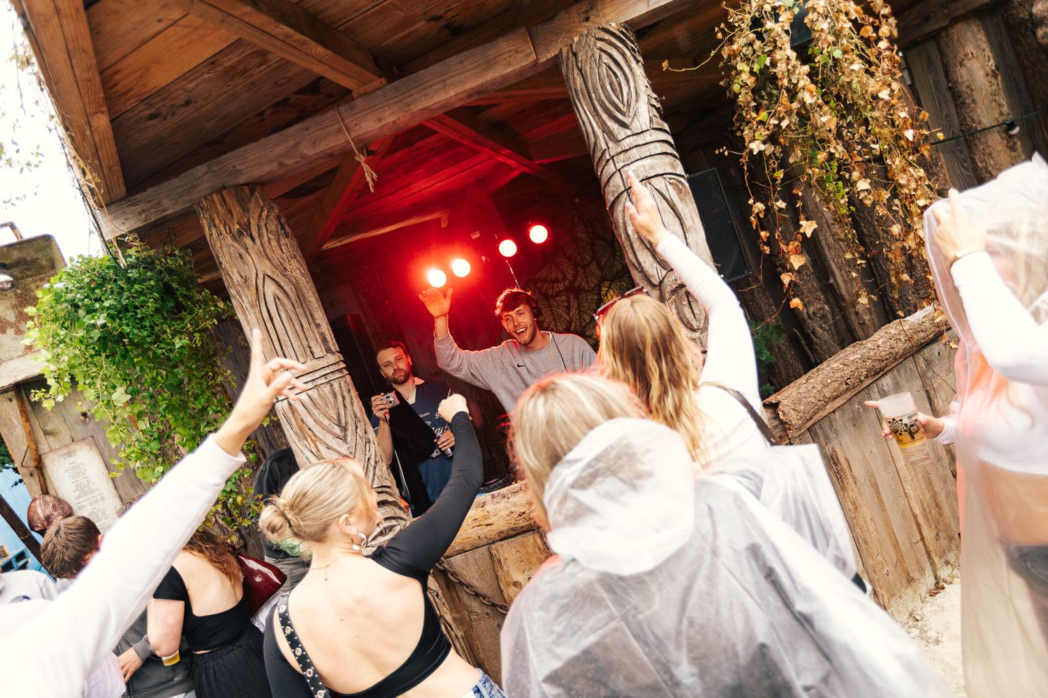 A lively wedding reception party scene with partygoers dancing under red lighting and rustic wooden beams.