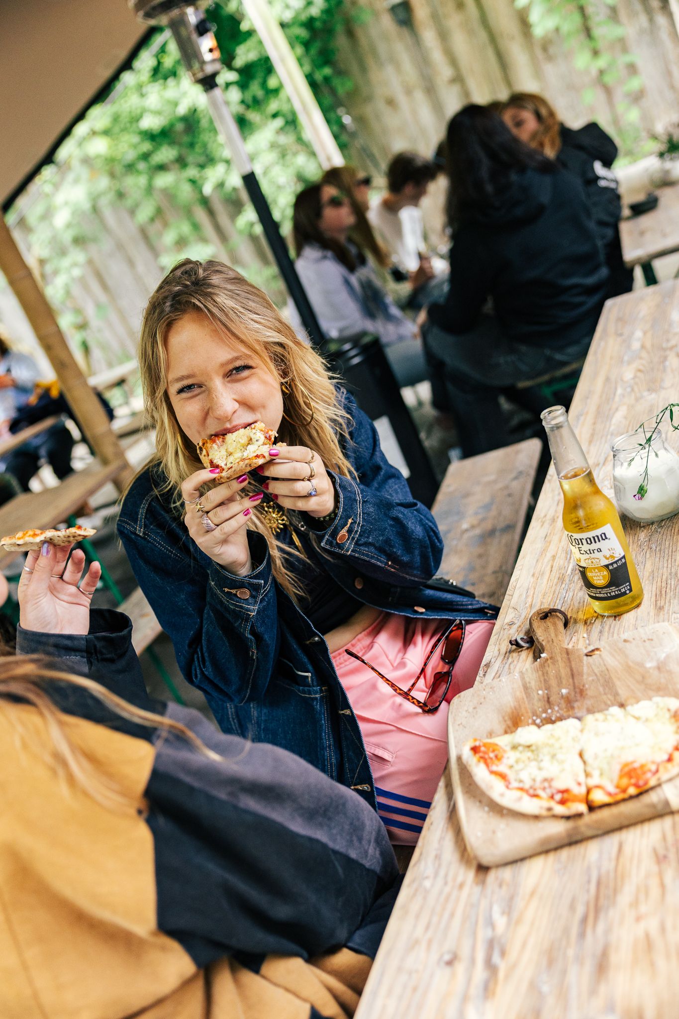A person in a navy jacket and pink shirt sits at a wooden table enjoying food and drinks in an outdoor dining setting.