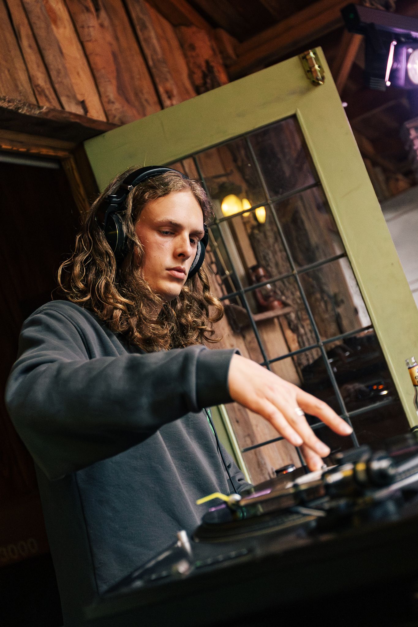 Someone with long wavy hair performs as a DJ using turntables in a dimly lit wooden interior space.