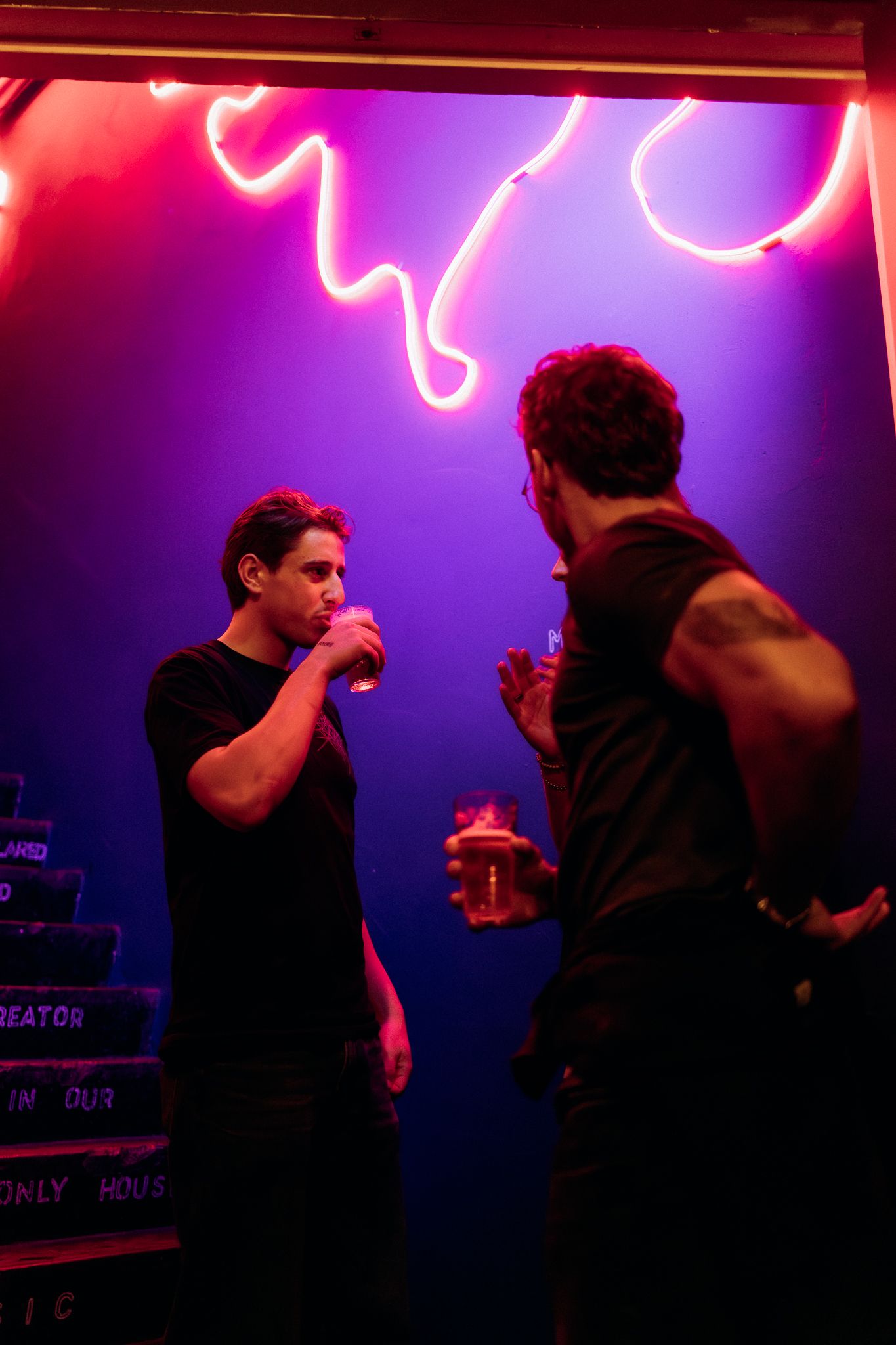 Silhouetted figures stand against a vibrant neon-lit backdrop in a nightclub atmosphere with red and purple lighting.