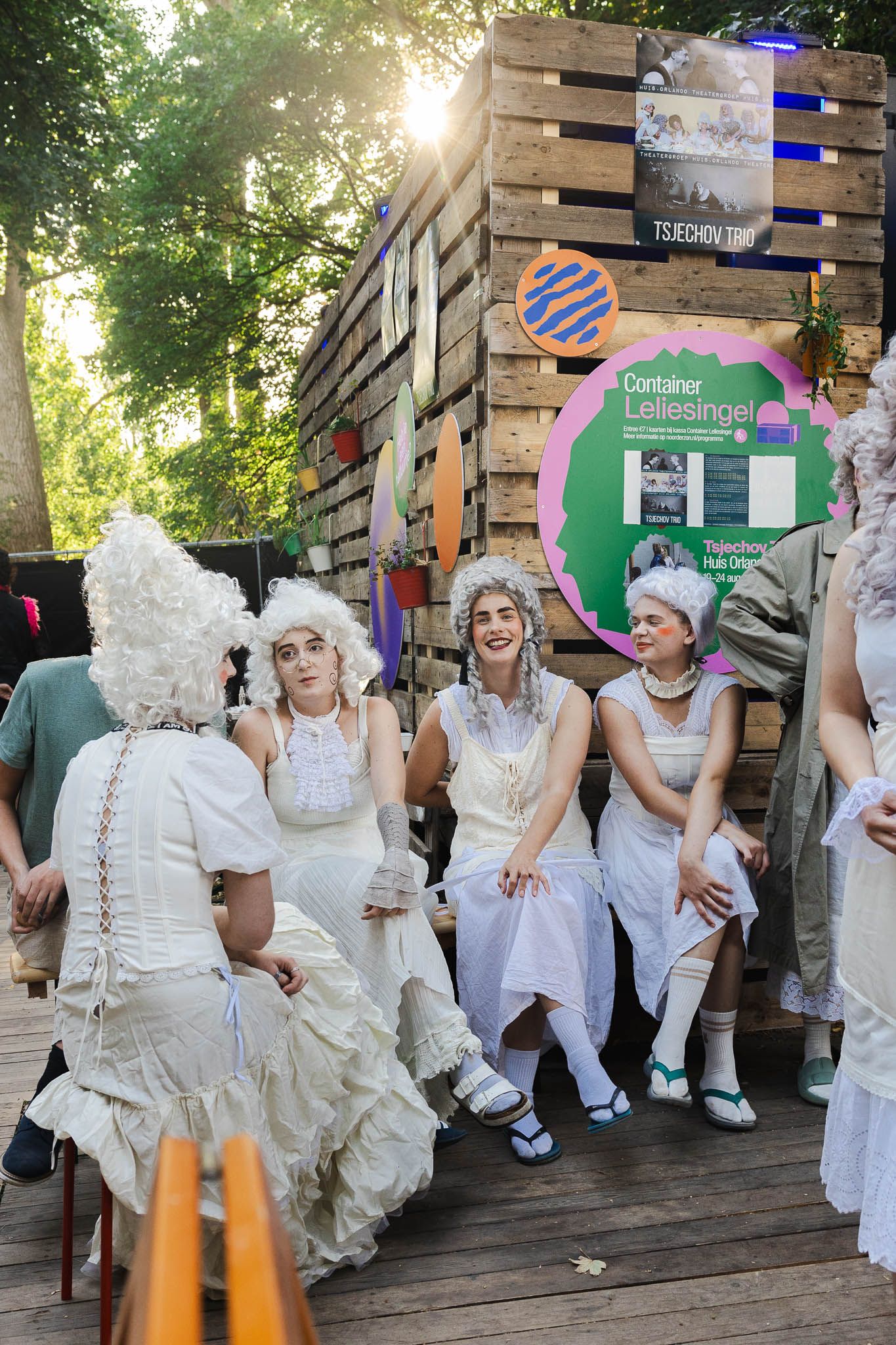 A group of performers dressed in all-white period costumes sit together on wooden steps during an outdoor event.