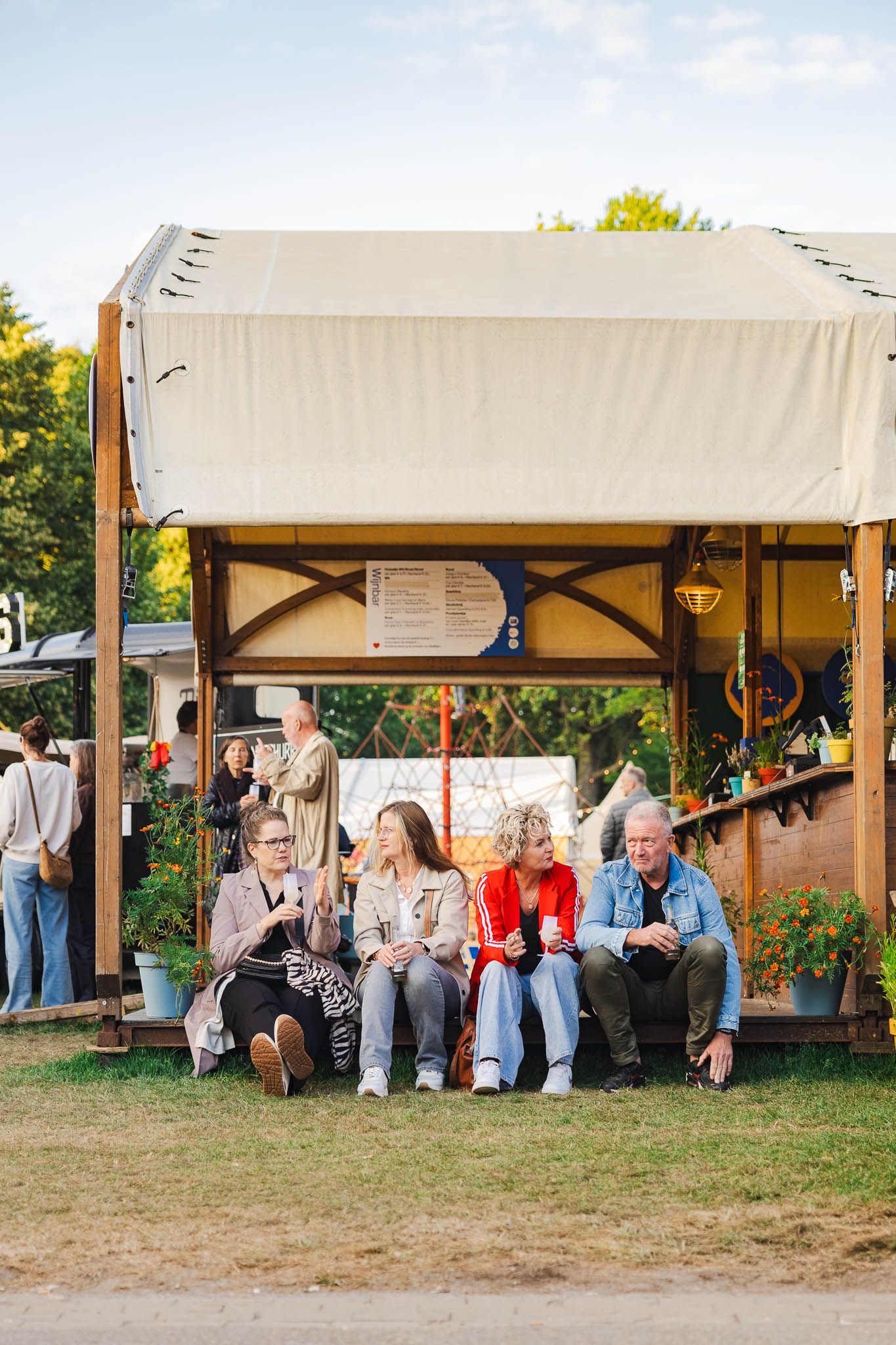 People relaxing under a white canopy tent at an outdoor festival or gathering during daytime.