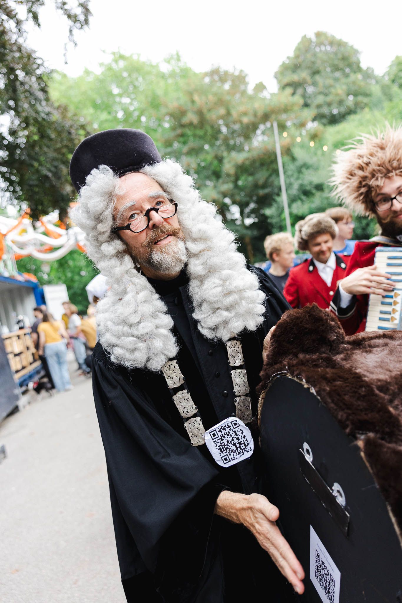 Man in black robe, white curly wig, and glasses at outdoor festival, resembling a judge or historical figure.
