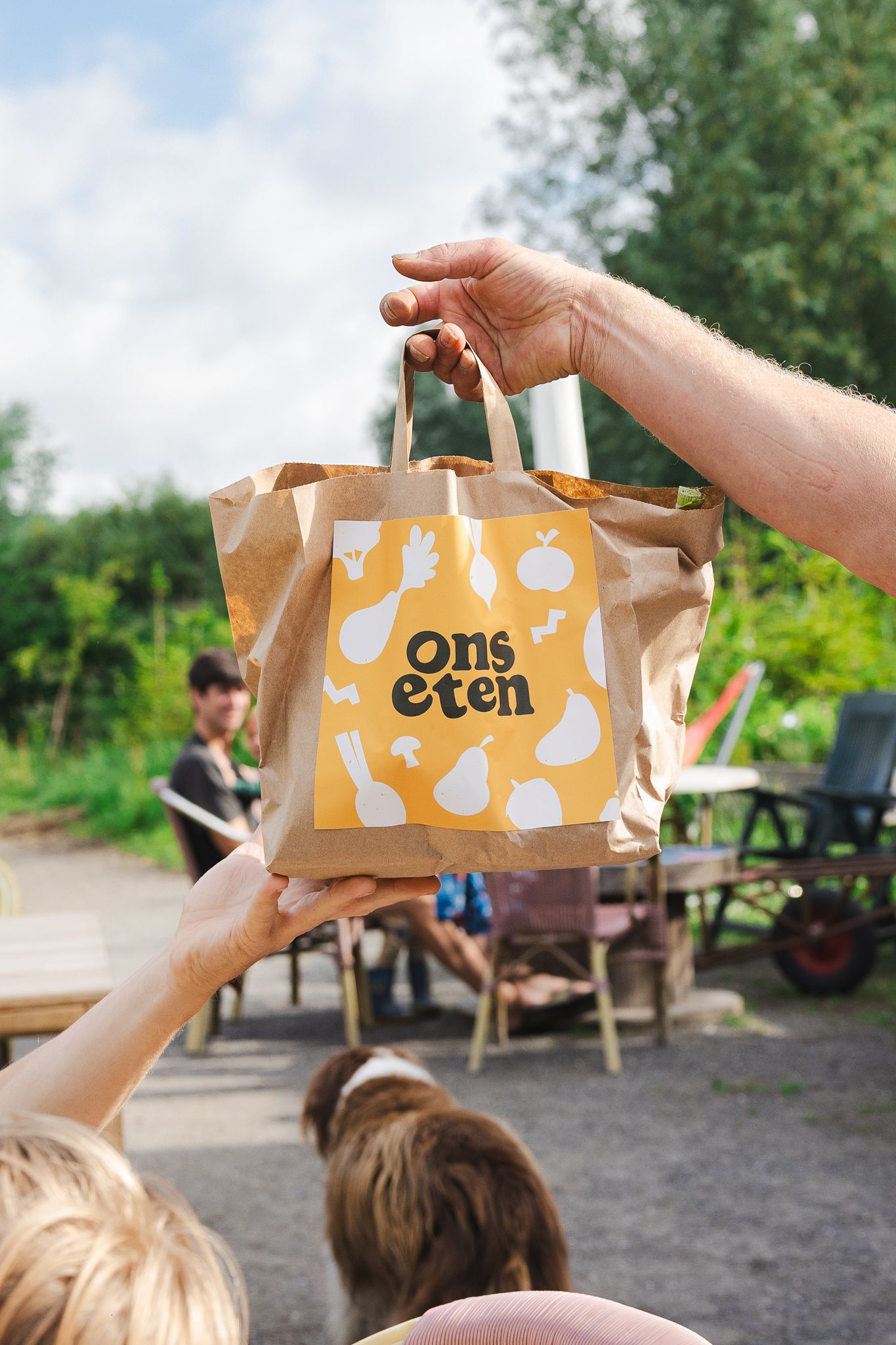 Hands passing a brown paper shopping bag with 'ons eten' text and musical note designs in an outdoor setting.