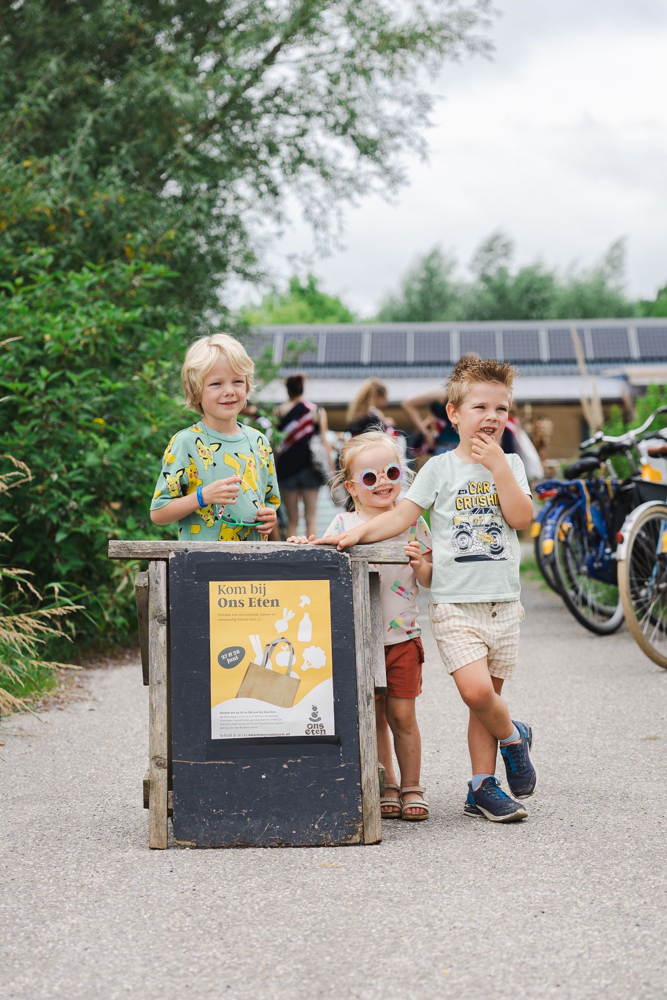 Two children play with a wooden toy box on a gravel path near a bike rack during an outdoor activity.
