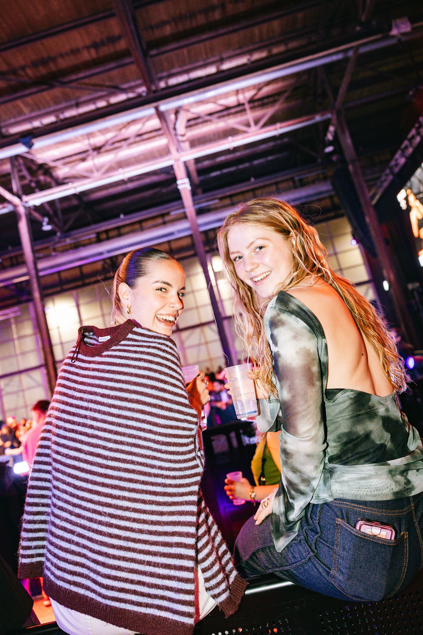 Two women smiling and holding drinks at an indoor event venue with purple lighting.
