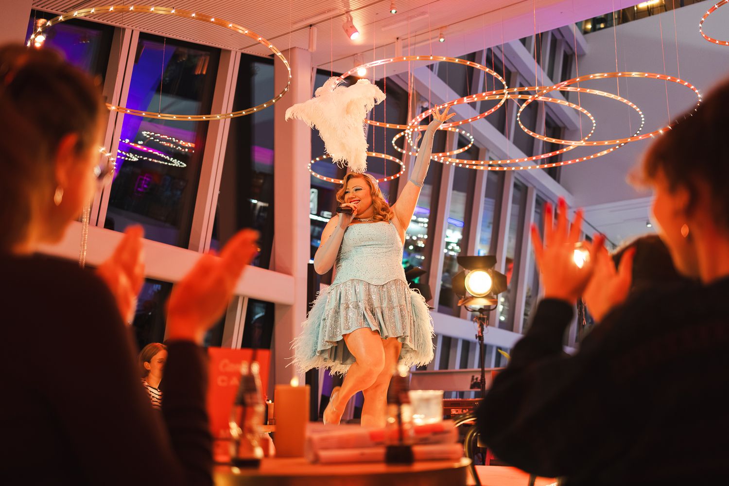 A woman in a light blue dress performs on a table at an upscale restaurant while guests applaud around her.