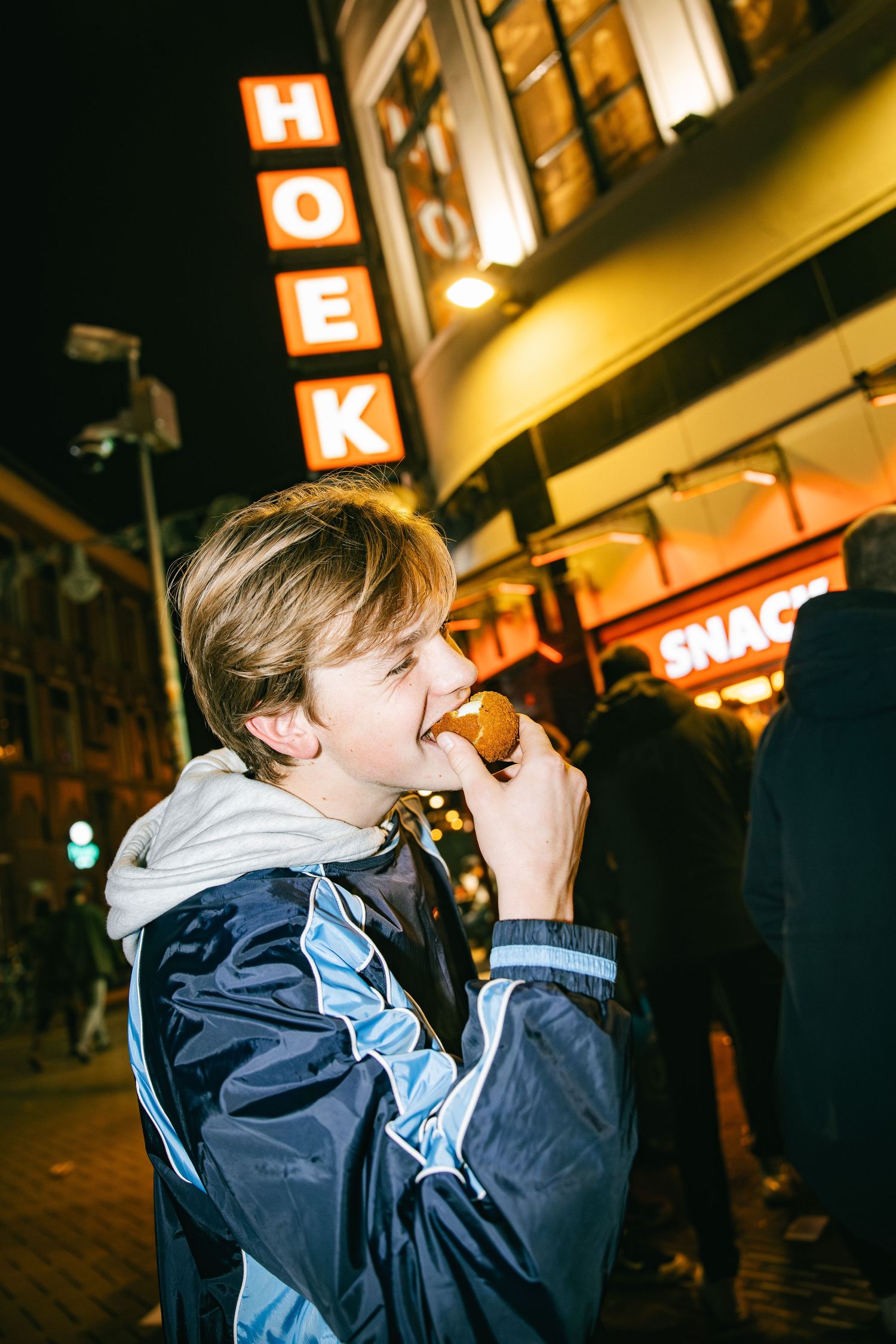 A person in a navy blue jacket drinks from a bottle at night with illuminated signs visible in the background.