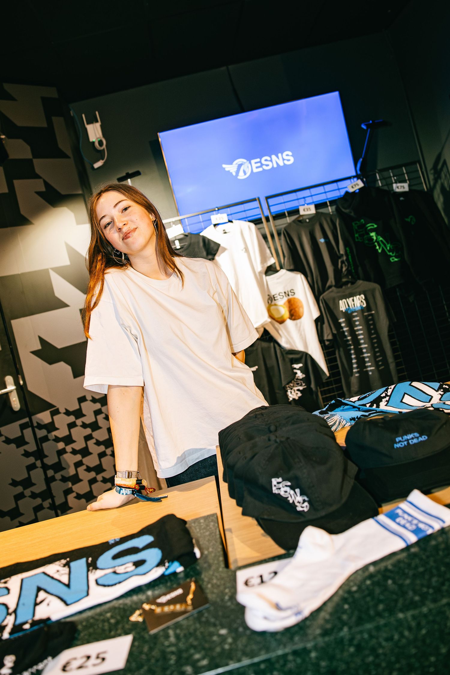 Person in white t-shirt working at a dark retail counter with merchandise displays visible in the background.