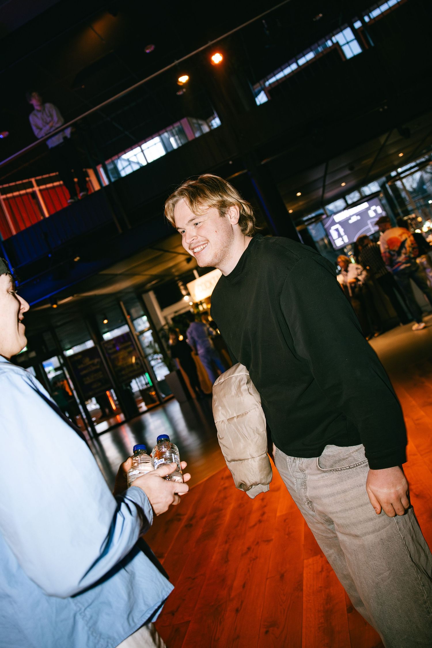 Two people interact in a dimly lit indoor space with wooden flooring and ambient lighting in the background.