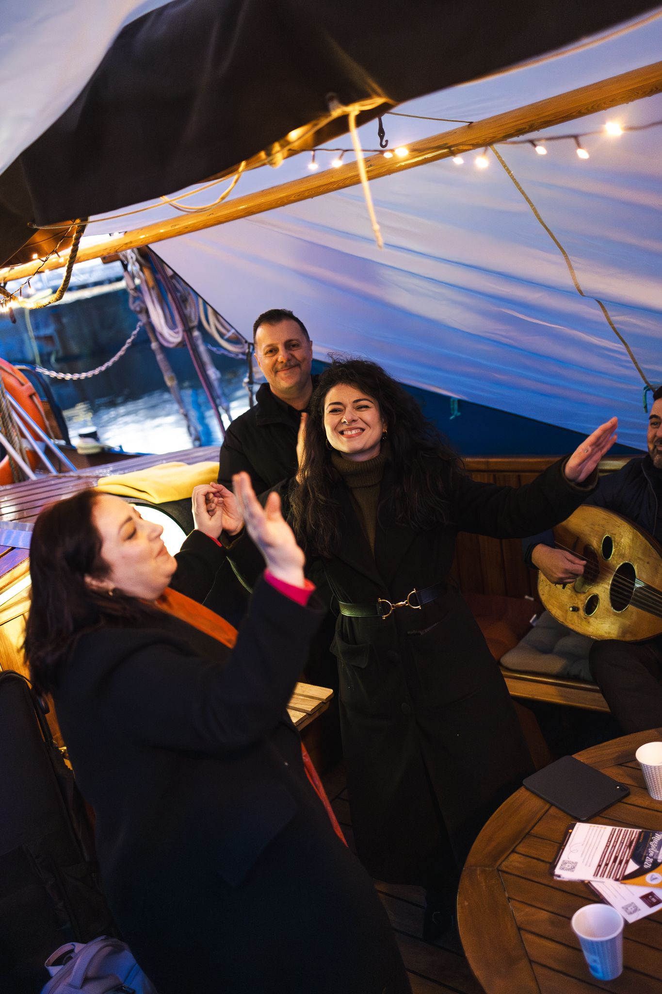 People enjoying a social gathering on a boat with a covered deck and wooden table at dusk.