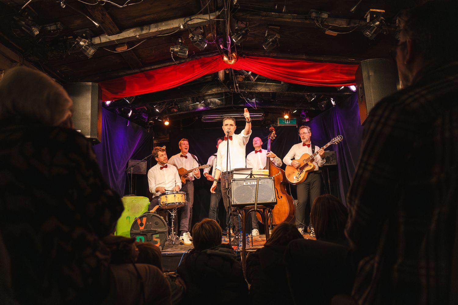 Musicians perform on stage with instruments under red and purple lighting at a live music venue.