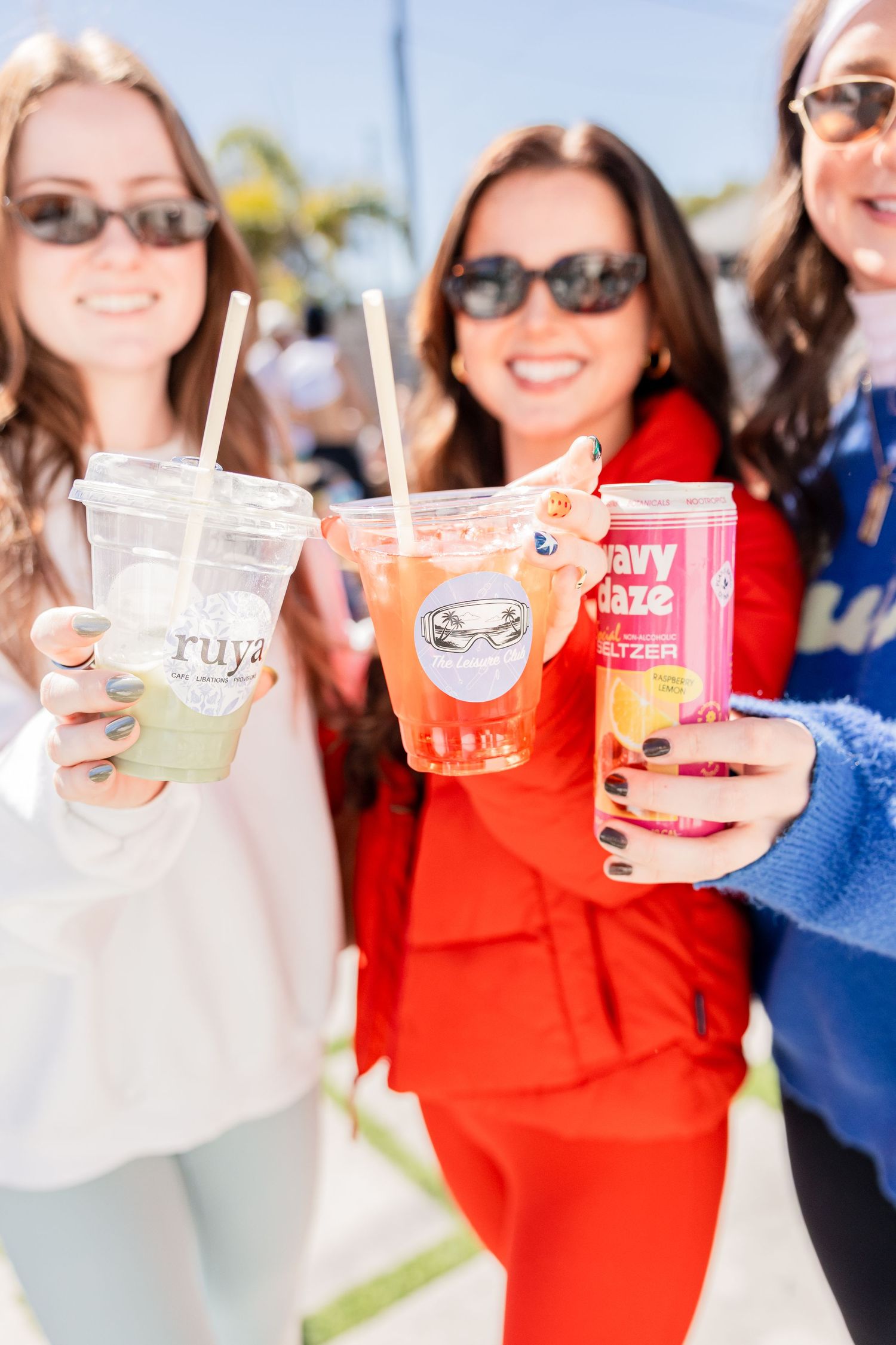 Three smiling women holding colorful drinks including a Wavy Daze seltzer at an outdoor event.