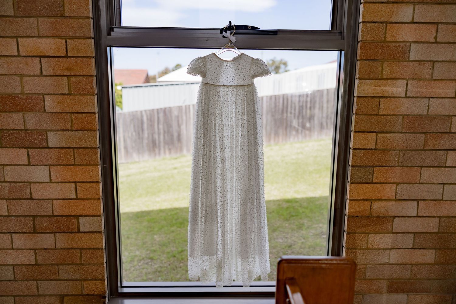 White christening gown hanging in window frame against brick wall backdrop.
