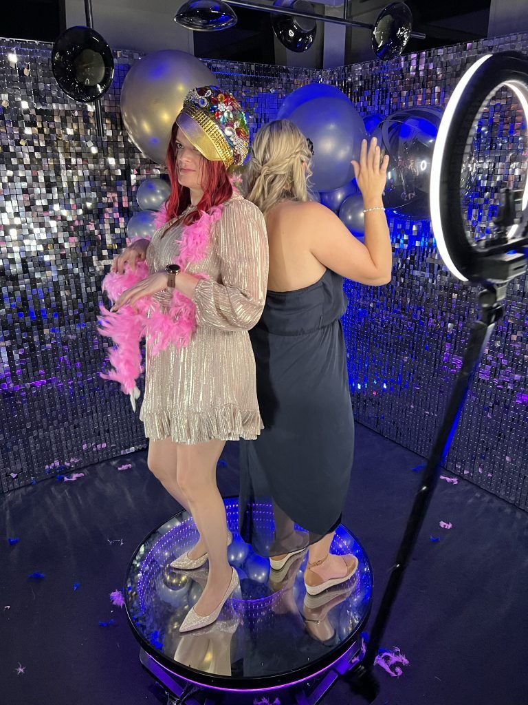 Two women posing at a brightly lit selfie station or ring light setup during a party event. 360 video booth vivitu venlo