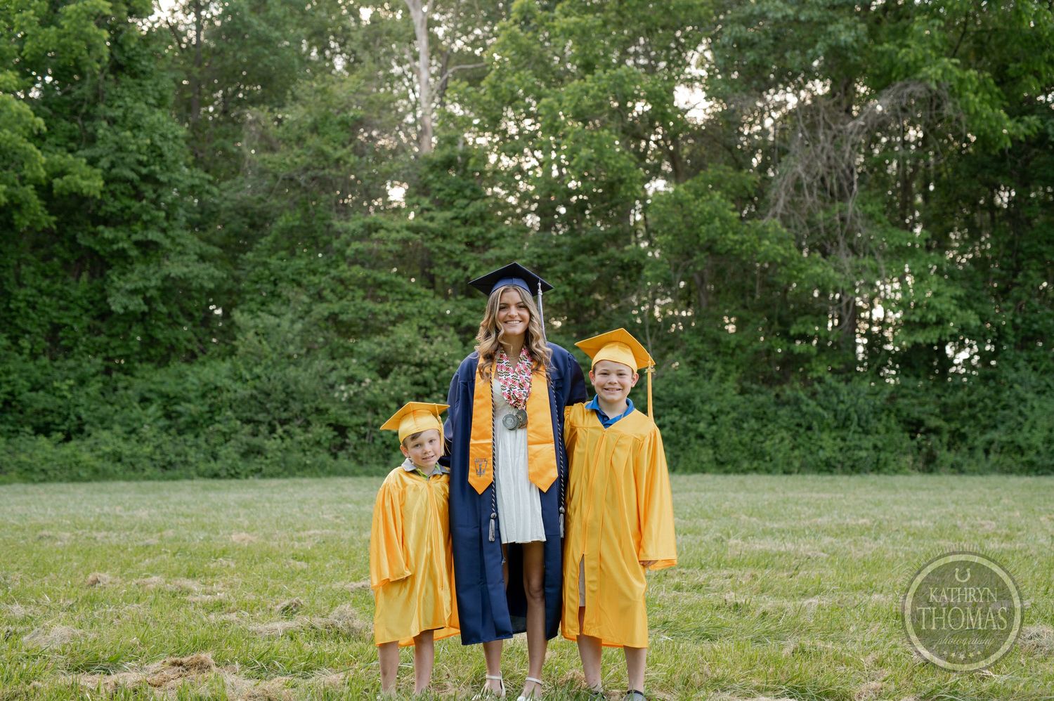 Three graduates in yellow and blue regalia pose together on grassy field.
