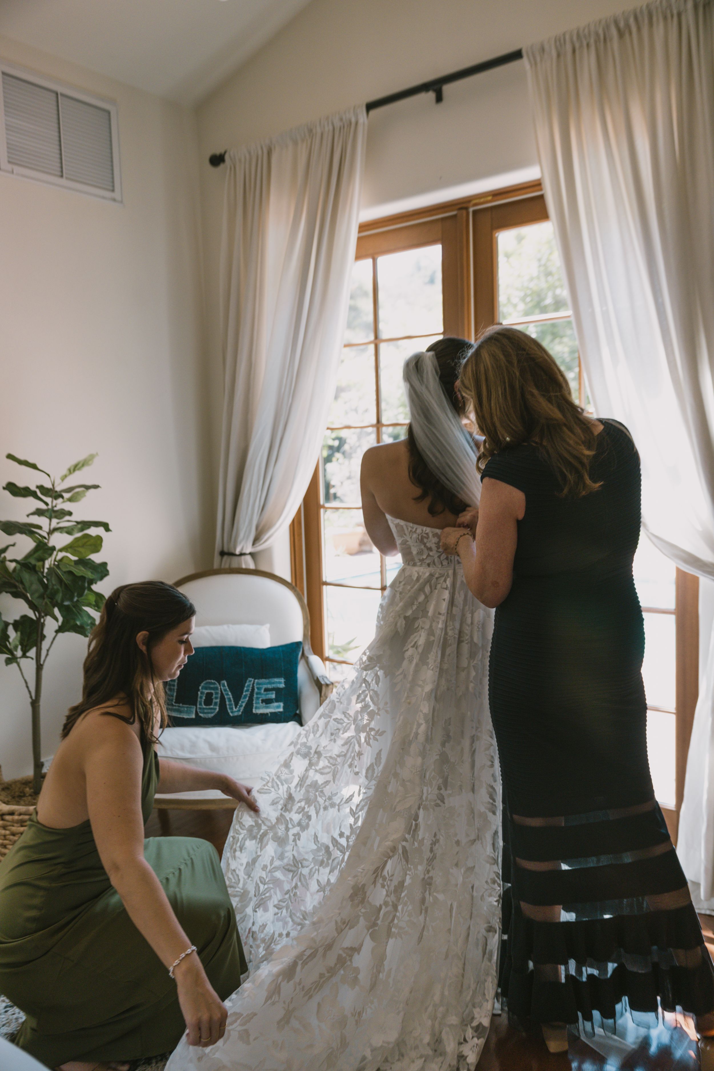 People helping to prepare and steam a white wedding dress hanging in a bright room.