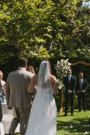 Wedding ceremony taking place outdoors under trees with wedding party lined up.